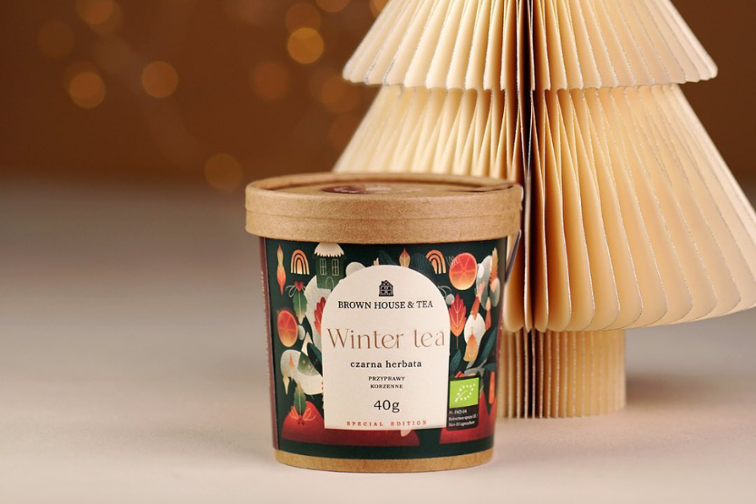 Puszka kraftowej herbaty Winter Tea od Brown House & Tea na tle papierowej choinki i ciepłego światła – świąteczna edycja czarnej herbaty